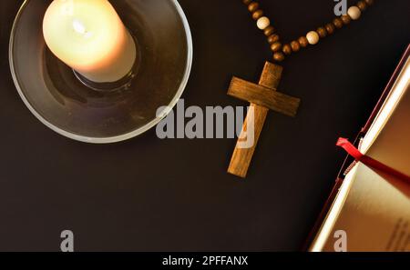 Schwarzer Nachttisch mit religiösen Elementen zum Beten mit einem christlichen Kreuz, einer offenen Bibel und einer beleuchteten Kerze auf einem Glasteller. Draufsicht. Stockfoto