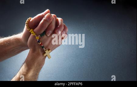 Christian betet mit Händen zusammen, mit Fingern, die mit Rosenkranzperlen verbunden sind, die mit isoliertem dunklem Hintergrund hängen. Erhöhte Ansicht von oben. Stockfoto