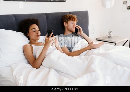Ein junges, wunderschönes gemischtrassiges Paar, das seine Handys hält und benutzt, während es zusammen im Bett liegt. Ein hübscher rothaariger, ernster Kerl, der telefoniert und seinen Bär Stockfoto