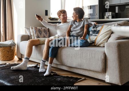 Ein junger, attraktiver, rothaariger, lächelnder Mann, der seine wunderschöne afrikanische Freundin ansieht und mit der Fernbedienung den fernsehsender wechselt, während sie sitzen, um abzuholen Stockfoto