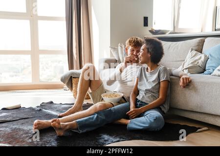 Junger, attraktiver Rothaariger, der seine afrikanische Freundin mit Popcorn fütterte, während sie auf dem Boden saßen und sich auf der Couch in einem gemütlichen, sonnigen Wohnzimmer zu Hause lehnten Stockfoto