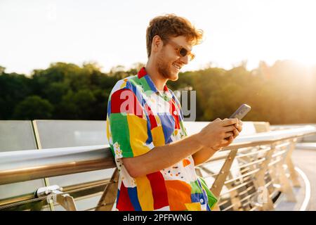 Ein junger, attraktiver, lächelnder Mann in einer Sonnenbrille, der sein Telefon hält und benutzt, während er in der Sonne auf dem Geländer steht Stockfoto