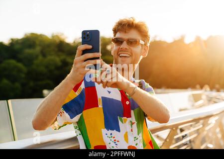 Ein junger, attraktiver, stylischer, lächelnder Mann mit Sonnenbrille, der Fotos auf seinem Handy macht, während er in der Sonne auf dem Geländer steht Stockfoto
