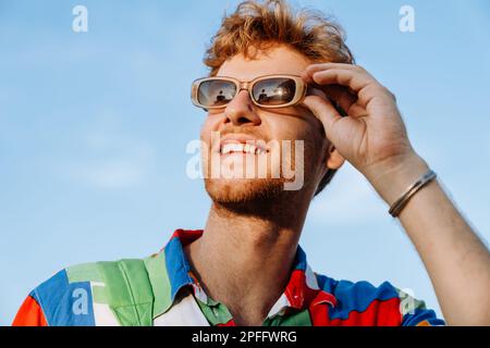 Porträt eines jungen, attraktiven, stylischen, lächelnden Mannes in einem farbenfrohen Hemd, der seine Sonnenbrille anpasst und zur Seite schaut Stockfoto