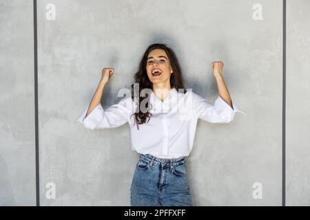 froh, dass Frau Pumpen Fäuste frohlockt ekstatischen feiert Erfolge auf graue Wand Hintergrund isoliert Stockfoto