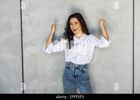 froh, dass Frau Pumpen Fäuste frohlockt ekstatischen feiert Erfolge auf graue Wand Hintergrund isoliert Stockfoto