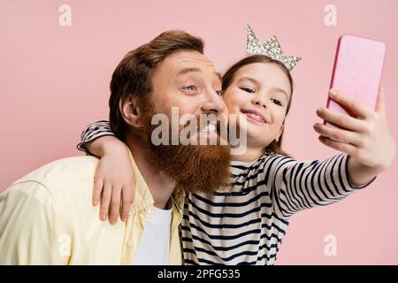Lächelndes Mädchen mit Kronenkopfband umarmt bärtigen Vater, während Selfie isoliert auf pinkfarbenem Stockbild gemacht wird Stockfoto