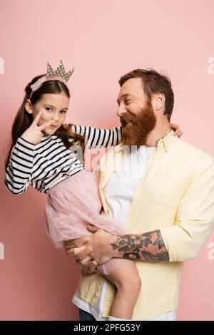 Ein überfreulicher Mann hält eine junge Tochter mit einem Kronenband, das ein Friedenszeichen auf pinkem Hintergrund zeigt, ein Stockbild Stockfoto