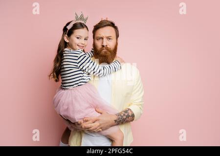 Lächelndes junges Mädchen mit Kronenband, das Kaninchenohren in der Nähe des Kopfes eines ernsthaften Vaters macht, auf rosa Hintergrund, Stockbild Stockfoto