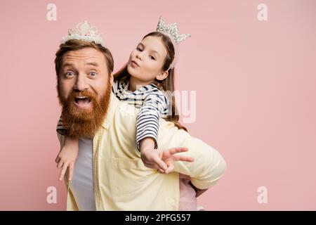 Ein Kind mit Kronenband zeigt ein Friedenszeichen, während er sich an Dad ranmacht, isoliert auf einem pinken, Stock-Bild Stockfoto