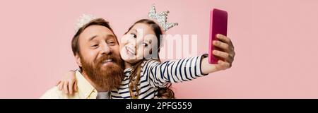 Positives Kind umarmt Vater mit Kronenband, während Selfie isoliert auf pinkfarbenem, bannerförmigem Stockbild gemacht wird Stockfoto