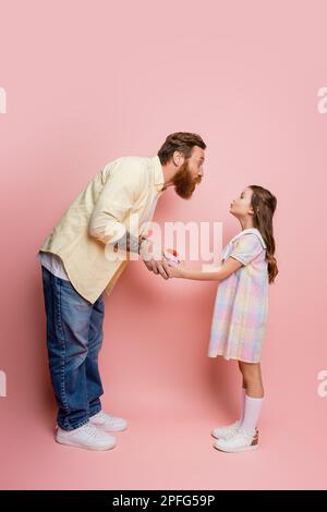 Seitenansicht eines Mannes, der die Lippen schmollt, während er der Tochter eine Geschenkbox überreicht, auf pinkfarbenem Hintergrund, Stockbild Stockfoto