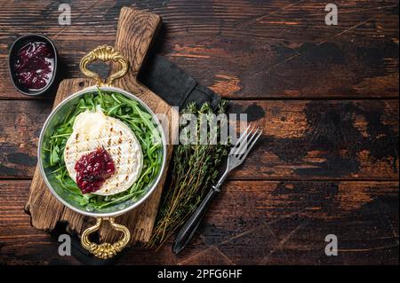 Gebackener Camembert Brie-Käse mit Preiselbeersauce und garniert mit Rucola-Salat in einer Pfanne. Holzhintergrund. Draufsicht. Speicherplatz kopieren. Stockfoto