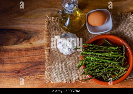 Draufsicht auf wilden Spargel in Tontopf auf einem Leintuch mit Eiern, Olivenöl und Knoblauch Stockfoto