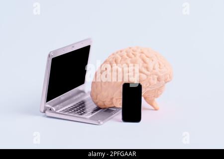 Detailliertes menschliches Hirnmodell mit einem Miniatur-Laptop und Smartphone, isoliert auf neutralem Hintergrund. Kreative Datenanalyse und künstliche Intelligenz Stockfoto