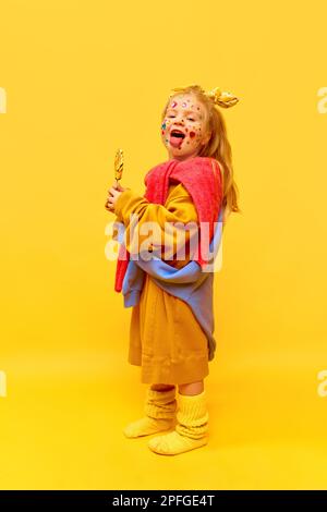 Foto des lustigen kleinen Mädchens mit Lutscher, die Zunge zeigt, große Kleidung trägt, vor der Kamera lächelt und einen gelben Hintergrund hat Stockfoto