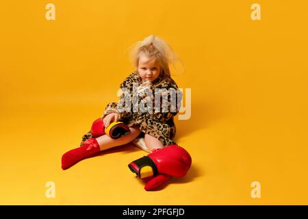 Ein kleines wütendes blondes Mädchen, das Boxhandschuhe und Leopardenmantel trägt, sitzt da und schaut weg über den gelben Hintergrund Stockfoto