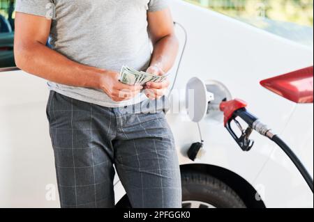 Ein Mann, der Geld zählt, das er für Benzin ausgeben muss. Konzept für Transportkosten. Zugeschnittene Ansicht eines Mannes, der Dollars im Hintergrund des Betankens eines Autos zählt. Stockfoto