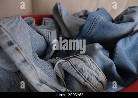 Gekrüppelte Jeanshosen, entfaltete Jeanshosen in einem roten Korb auf einem weißen Sofa. Gekrüppelte Denimhosen Konzeptfoto. Vorderansicht der sauberen Denimhose. Stockfoto