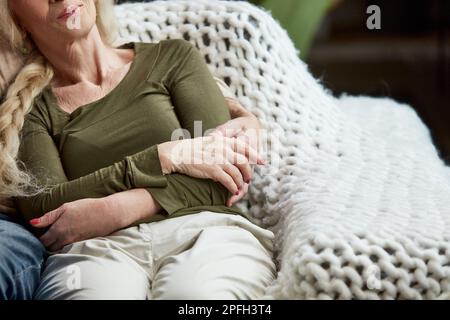 Zugeschnittenes Bild einer Seniorin, Frau, die mit ihrem Mann auf der Couch sitzt und sie umarmt, unterstützt und aufmerksam macht. Gemütlichkeit Stockfoto