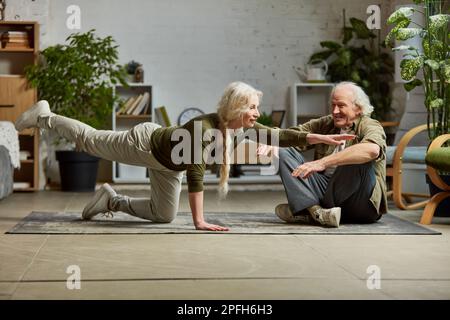 Reife Frau mittleren Alters, die mit ihrem älteren Mann zu Hause Yoga-Übungen macht. Verbinden Sie Training, Unterstützung und Unterstützung Stockfoto