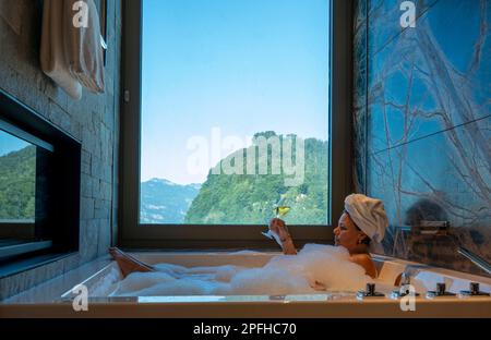 Frau liegt in einer Badewanne mit einem Glas Champagner und genießt den Fensterblick über den Berg an einem sonnigen Sommertag in Burgenstock, Nidwalden, Switzerlan Stockfoto