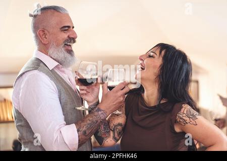 Zwei Hipster-Liebhaber mittleren Alters, ein Mann mit einem langen weißen Bart und Pferdeschwanz, und eine brünette Frau, die in einem verschwommenen Hintergrund steht und auf ihren Sieg anstoßen Stockfoto