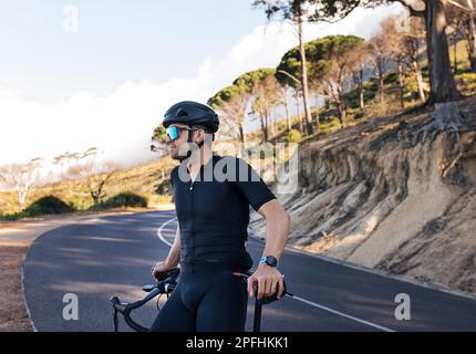 Seitenansicht eines professionellen Motorradfahrers, der sich auf seinem Road Bike entspannt. Männlicher Radfahrer mit Brille und Helm genießt die Aussicht und macht eine Pause während der Stockfoto