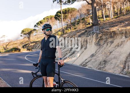 Porträt eines professionellen Radfahrers im Helm, der auf der Straße wegschaut. Sportler in schwarzer Sportbekleidung mit Roadbike, der sich auf dem Land entspannt Stockfoto