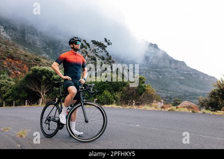 Junger Radfahrer, der sich nach dem Training mit den Händen auf der Hüfte entspannt. Profisportler mit Road Bike genießt die Aussicht, während er auf der Straße steht Stockfoto