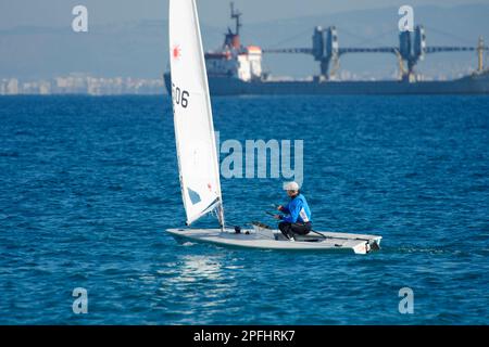 Foto eines Teenagers, der auf einem Laser-Segelboot mit einem riesigen Schiff im Hintergrund segelt Stockfoto