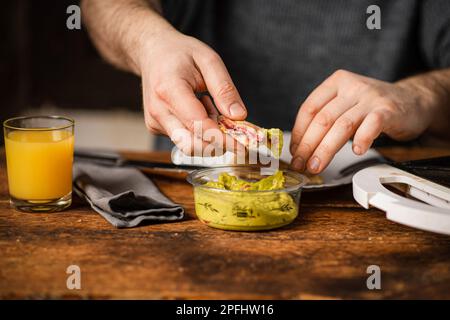 Ein unbekannter Mann isst Toast mit Guacamole. Frühstückszeit. Die Hände des Mannes halten einen Toast. Stockfoto