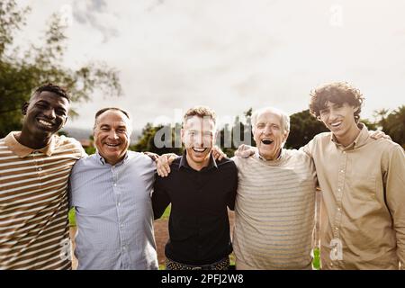 Glückliche, generationsübergreifende Gruppe von Männern mit unterschiedlichen Alters und ethnischen Zugehörigkeiten, die Spaß haben, vor der Kamera im Park zu lächeln Stockfoto