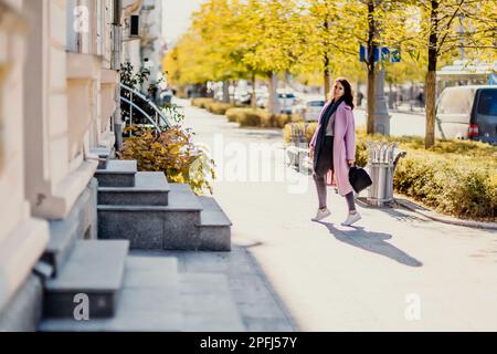 Frau Herbststadt. Eine Frau in einem rosa Kunstpelzmantel, die im Herbst an einem sonnigen Tag auf einer Straße posiert. Bäume mit gelben Blättern entlang der Straße. Stockfoto