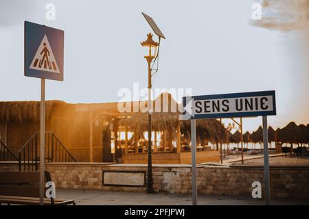 Einbahnschilder und Fußgängerüberquerung auf der Zufahrtsstraße zum Strand, im Sonnenaufgangslicht. Strohschirme sind im Hintergrund zu sehen Stockfoto