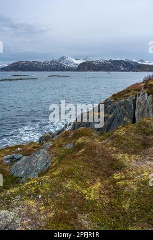 Stürmischer Ozean am Ufer von Hillesøya, in Troms, Norwegen, mit Wellen an der Küste kleiner Inseln, mit schneebedeckten Bergen im Hintergrund.