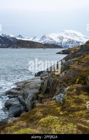 Stürmischer Ozean am Ufer von Hillesøya, in Troms, Norwegen, mit Wellen an der Küste kleiner Inseln, mit schneebedeckten Bergen im Hintergrund. Stockfoto