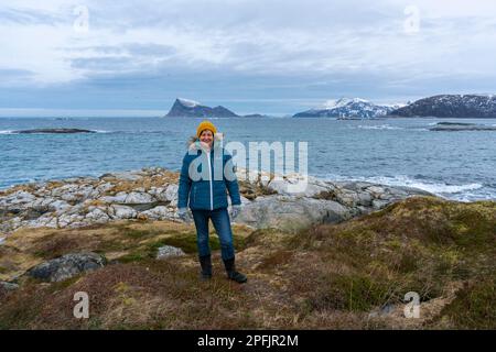 Glückliche Frau am Ufer des Nordatlantiks, an einem knappen Wintertag, ohne Schnee an der Küste. Stürmisches Wetter mit Inseln in Hillesøya, Norwegen Stockfoto