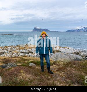 Glückliche Frau am Ufer des Nordatlantiks, an einem knappen Wintertag, ohne Schnee an der Küste. Stürmisches Wetter mit Inseln in Hillesøya, Norwegen Stockfoto