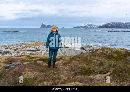 Glückliche Frau am Ufer des Nordatlantiks, an einem knappen Wintertag, ohne Schnee an der Küste. Stürmisches Wetter mit Inseln in Hillesøya, Norwegen Stockfoto