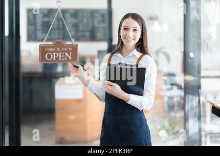 Eine junge asiatische Frau, die ein kleines Unternehmen eröffnete und ein lächelndes Gesicht auf einer Schürze vor einer Theke der Café-Bar zeigte. Unternehmen Stockfoto