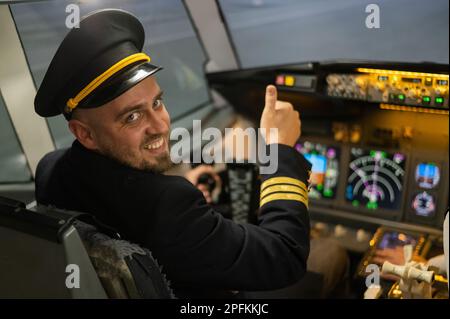 Kaukasischer bärtiger Mann, der beim Fahren eines Flugsimulators lächelt. Der Pilot im Cockpit zeigt Daumen nach oben. Stockfoto
