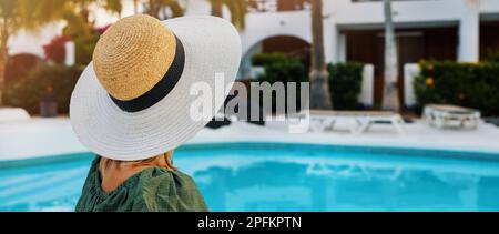 Eine nicht wiedererkennbare Frau mit Hut, die sich am Pool in einem luxuriösen Hotelresort erholt und einen perfekten Urlaub genießt. Banner mit Kopierbereich Stockfoto