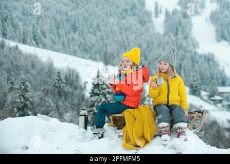 Kinder auf Picknick im Winter. Kleines Paar, das im Schnee auf einem Schlitten sitzt und heißen Tee trinkt, um die Weihnachtszeit zu genießen. Wintercamping. Stockfoto