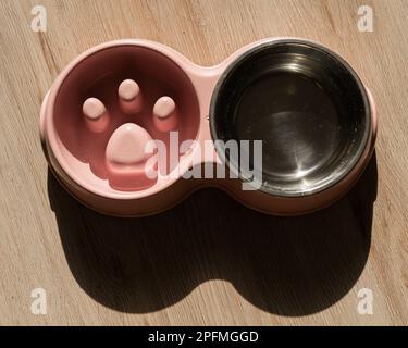 Eine Doppelschüssel für langsames Füttern und eine Schüssel Wasser für den Hund. Draufsicht auf einen rosafarbenen Teller mit trockenen Speisen auf einem Holzboden. Stockfoto