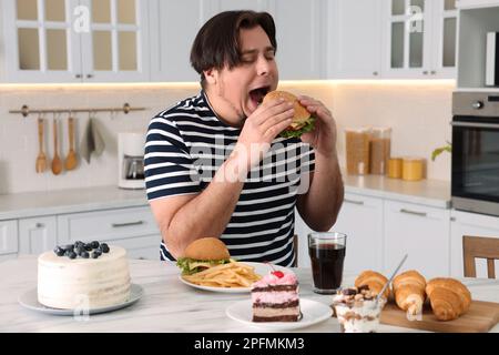 Hungriger, übergewichtiger Mann, der leckere Burger am Tisch in der Küche isst Stockfoto