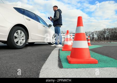 Ausbilder mit Klemmbrett in der Nähe des Fahrzeugs im Freien, konzentrieren Sie sich auf den Verkehrskegel. Fahrschulprüfung Stockfoto