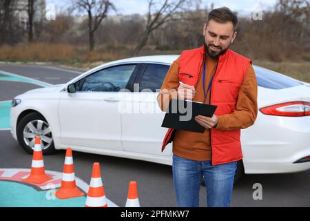 Kursleiter mit Klemmbrett in der Nähe des Fahrzeugs auf der Teststrecke. Fahrschule Stockfoto