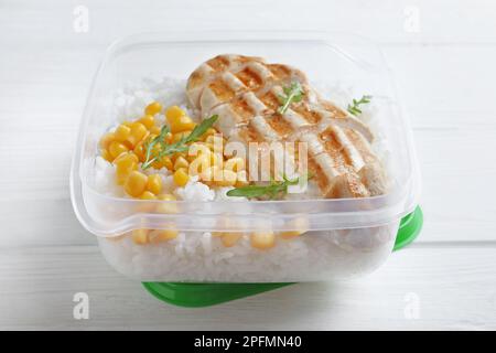 Schmackhafter Reis mit gegrilltem Fleisch und Mais in einem Kunststoffbehälter auf einem weißen Holztisch Stockfoto