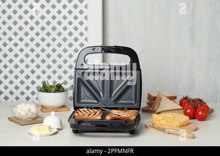 Moderne Grillmaschine mit Sandwiches und verschiedenen Produkten auf einem weißen Tisch Stockfoto